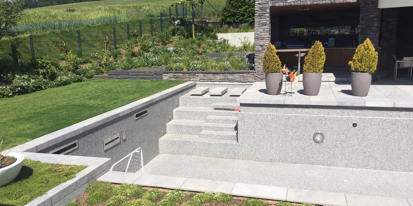 Leerer moderner Pool mit Treppen und grauen Fliesen, umgeben von grüner Wiese und Steinterrasse mit drei Pflanzenkübeln.