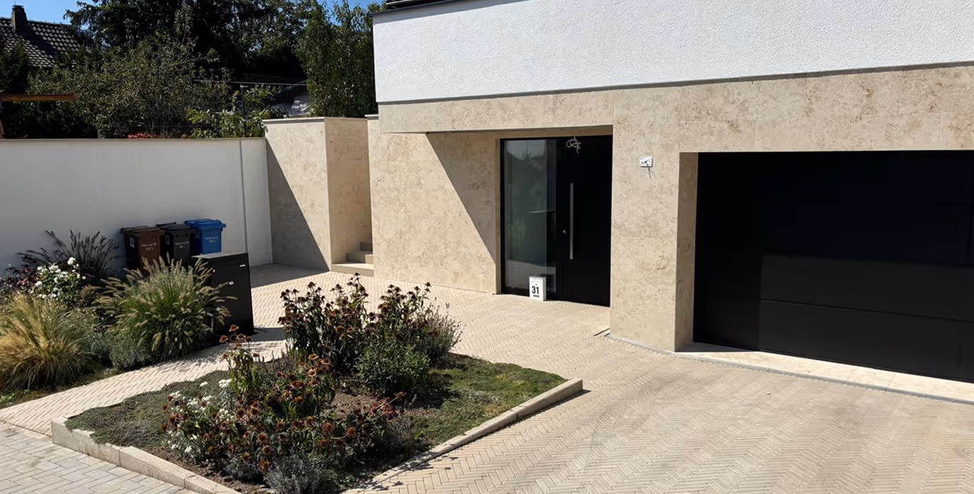 Moderne Hausfassade mit beigem Stein, schwarzer Eingangstür, schwarzer Garagentür und einem kleinen Garten mit Pflanzen und Blumen.
