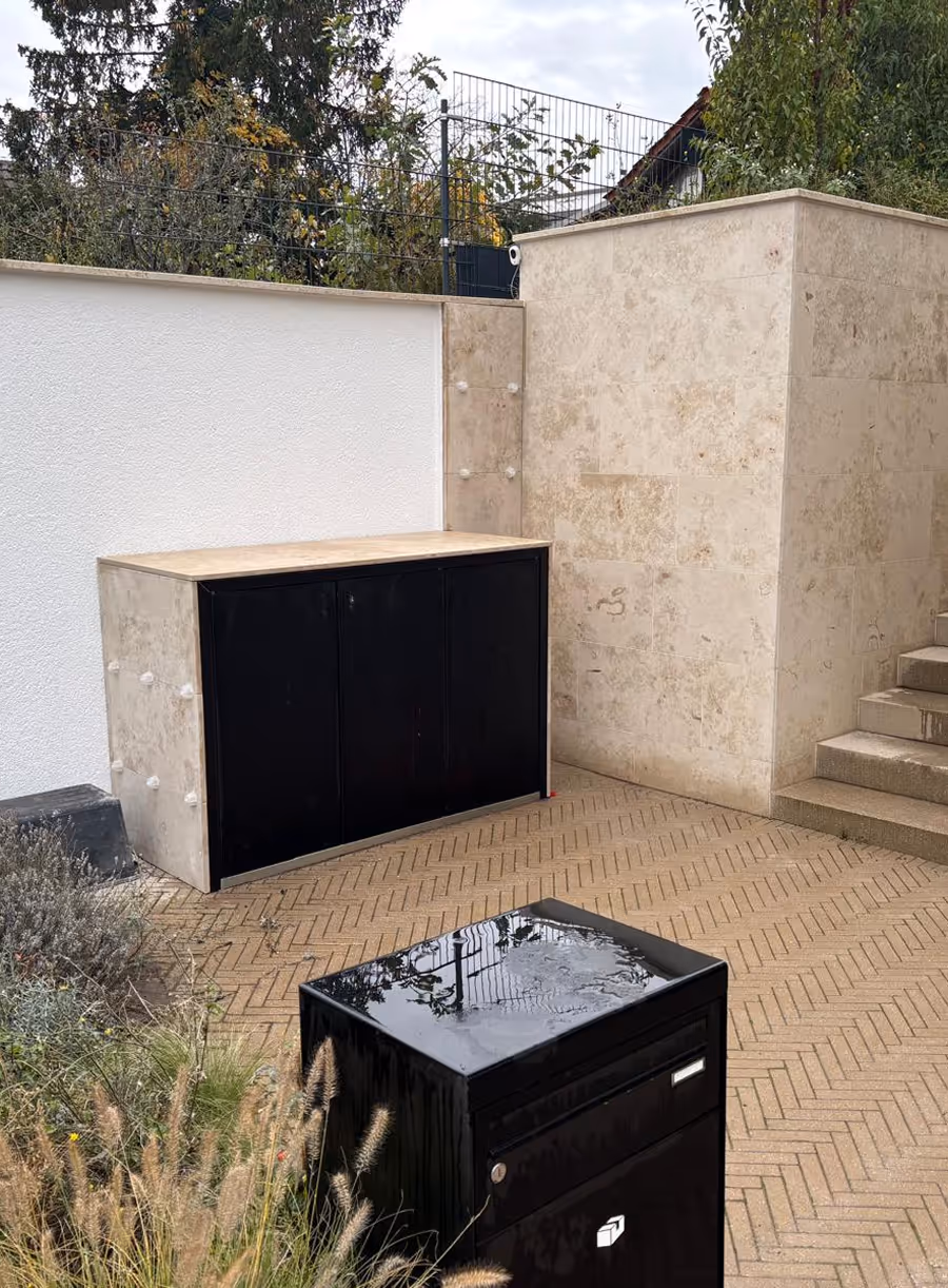 Moderner Außenbereich mit zwei schwarzen Metallbehältern auf beige gepflastertem Boden nahe einer hellen Steinwand und Treppe.