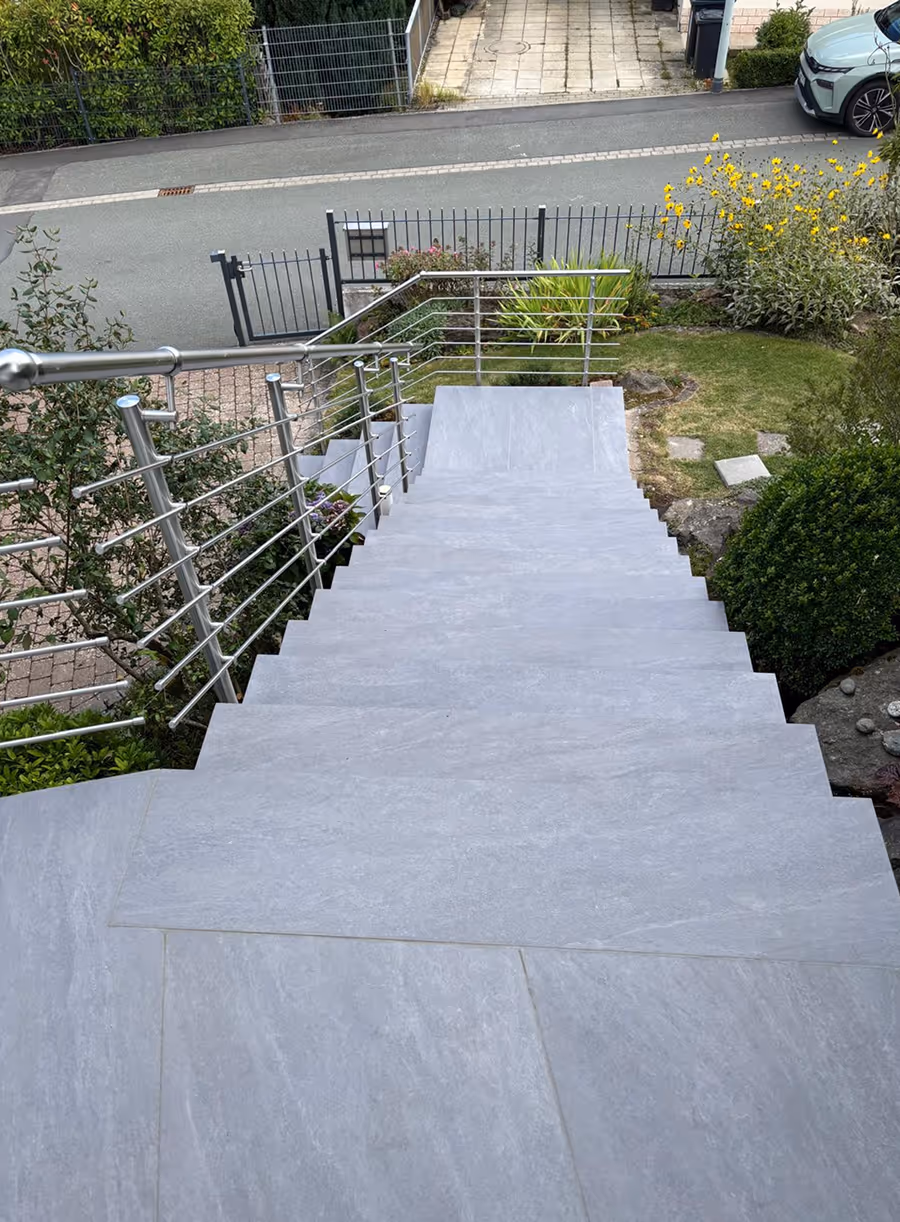 Blick nach unten auf eine außenliegende, graue Treppe mit Edelstahlgeländer, umgeben von Garten mit grünen Pflanzen und gelben Blumen, im Hintergrund eine graue Straße und ein Auto.