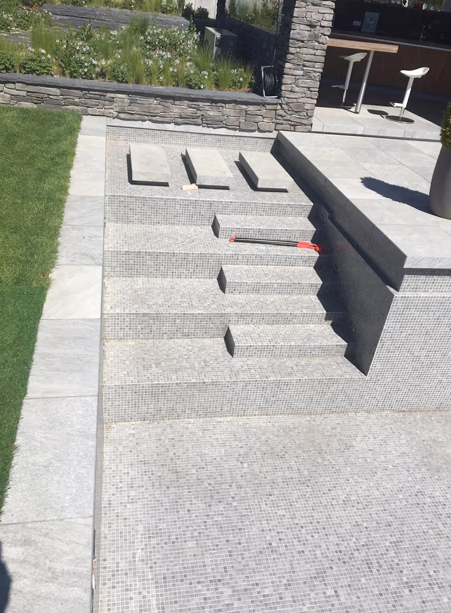 Leeres, modernes Schwimmbecken mit grauen Mosaikfliesen, Treppen und angrenzendem Garten mit Steinmauer und Sitzbereich.