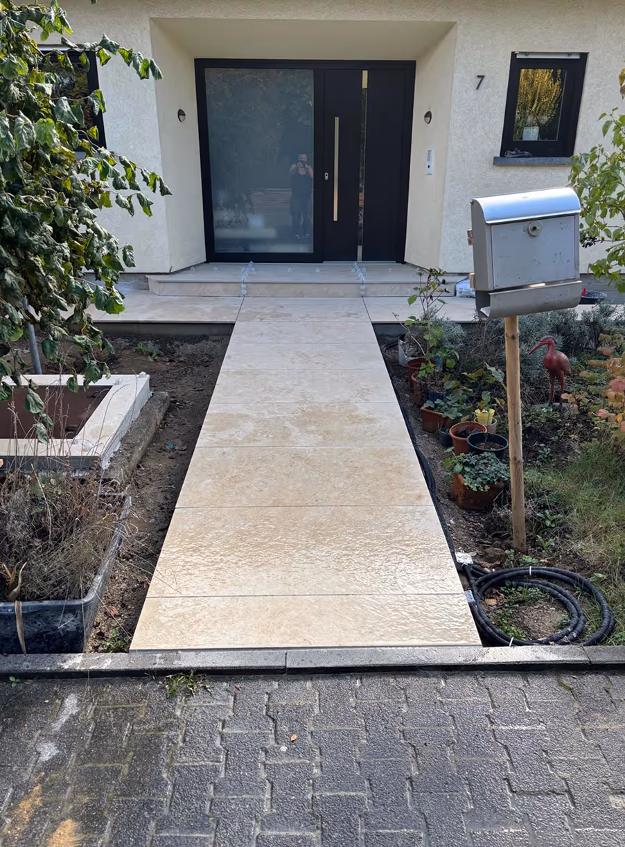 Heller Natursteinweg führt zu moderner Haustür mit schwarzem Rahmen, rechts Briefkasten auf Holzpfosten und Pflanzen.