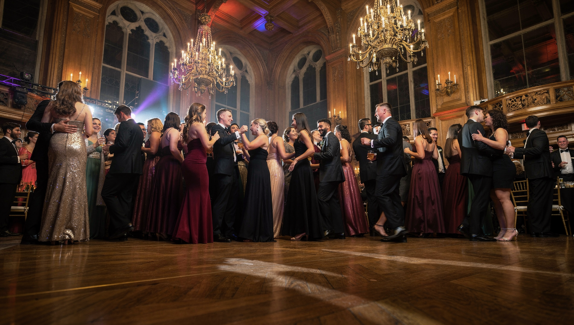 Guests dressed in formal gowns and tuxedos socialize and dance in a grand hall with chandeliers.