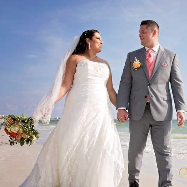 Bride in white dress and groom in gray suit holding hands and smiling on a beach.