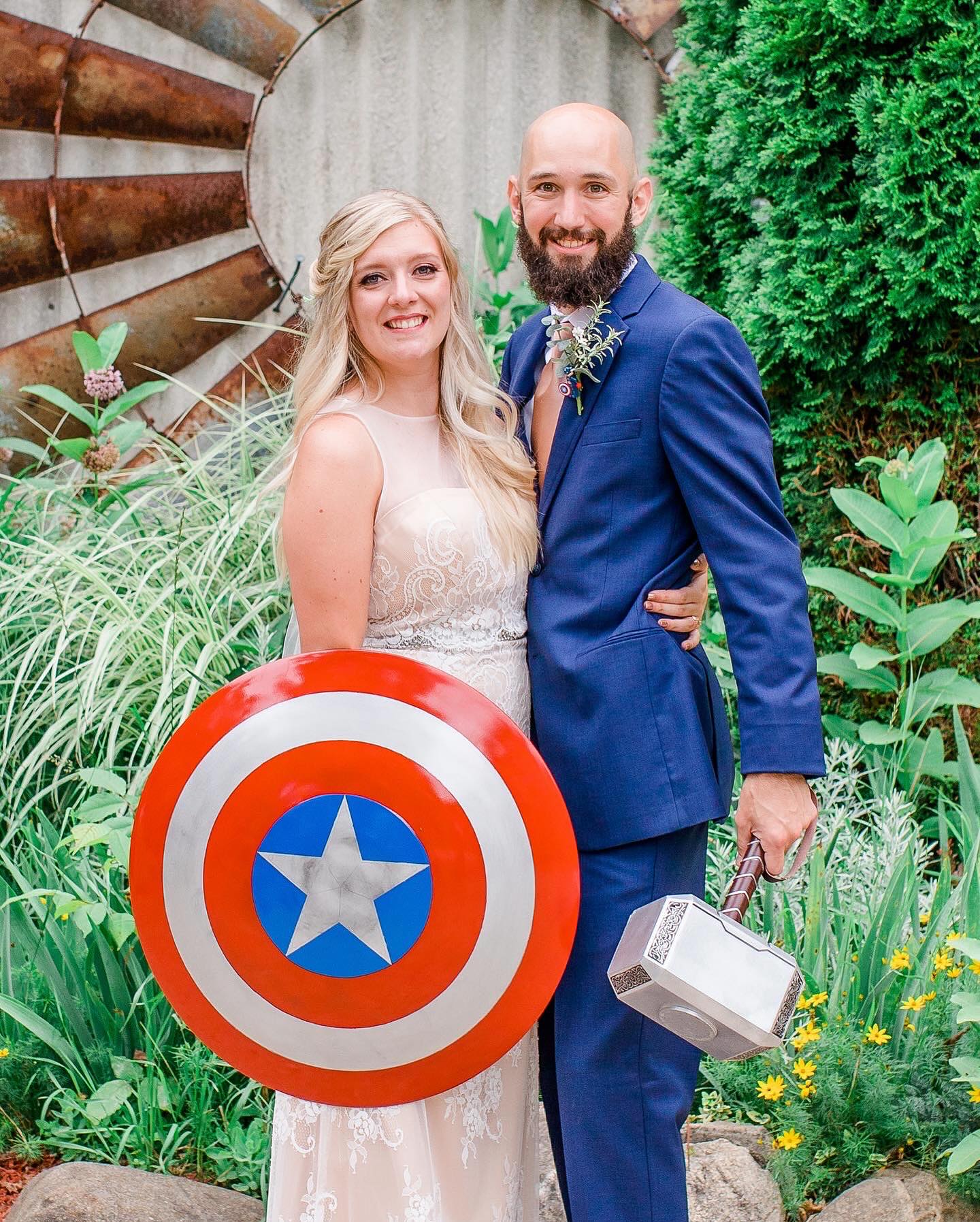 Bride in white dress holding Captain America shield and groom in blue suit holding Thor hammer smiling.