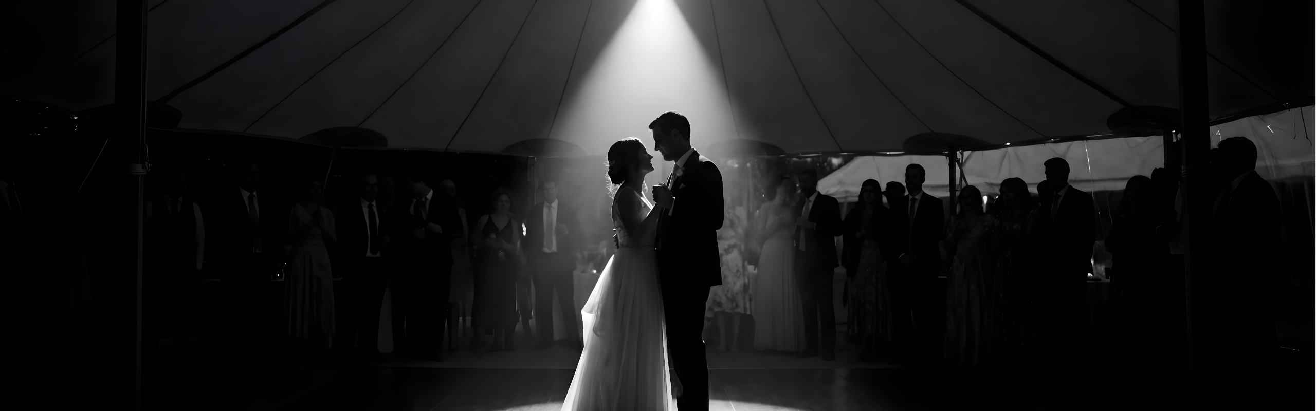 Bride and groom dancing under a spotlight surrounded by guests in a wedding tent.