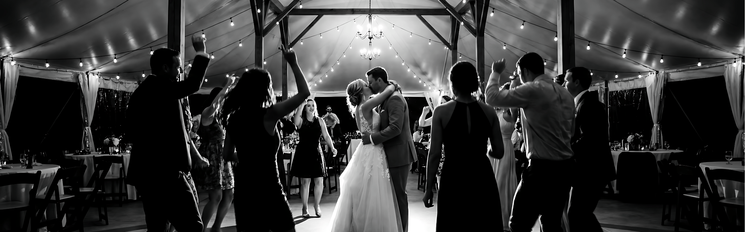 Bride and groom kissing on dance floor under string lights while guests dance around them in tent.