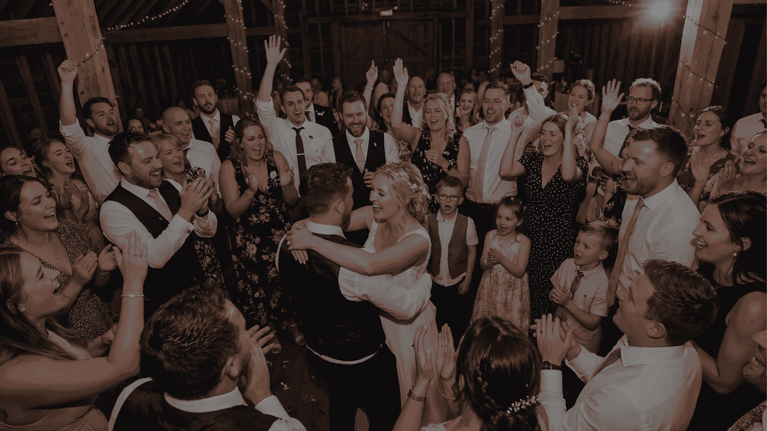 Bride and groom dance surrounded by cheering and clapping wedding guests in a warmly lit rustic barn.