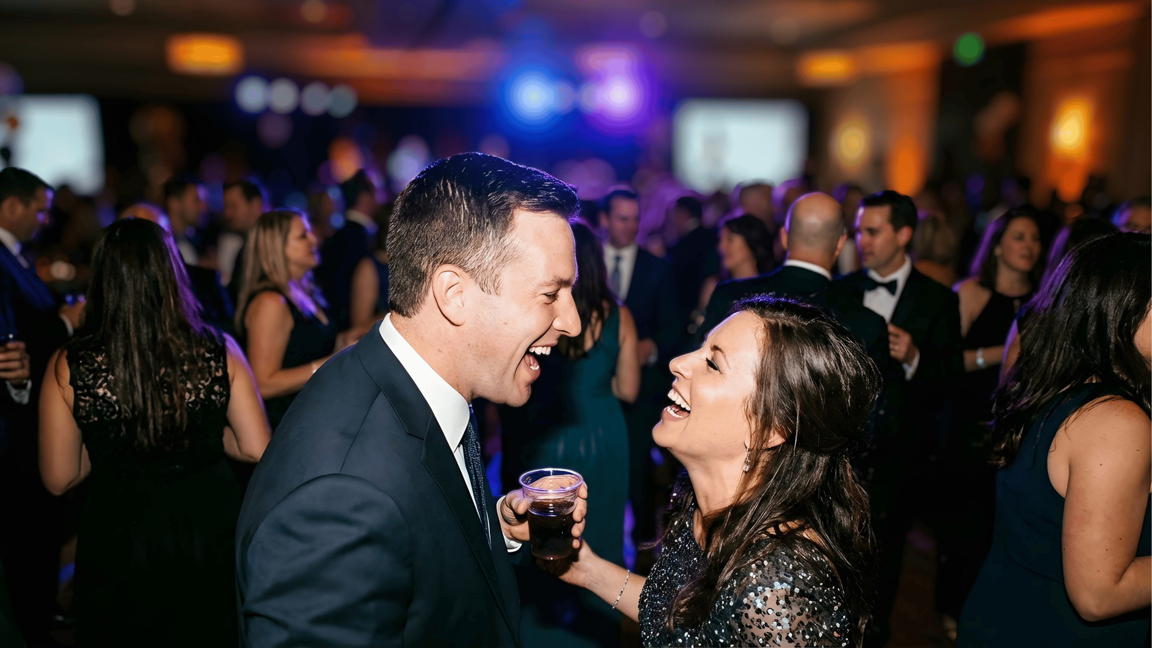 Man and woman laughing and holding drinks at a formal party with other guests in the background.