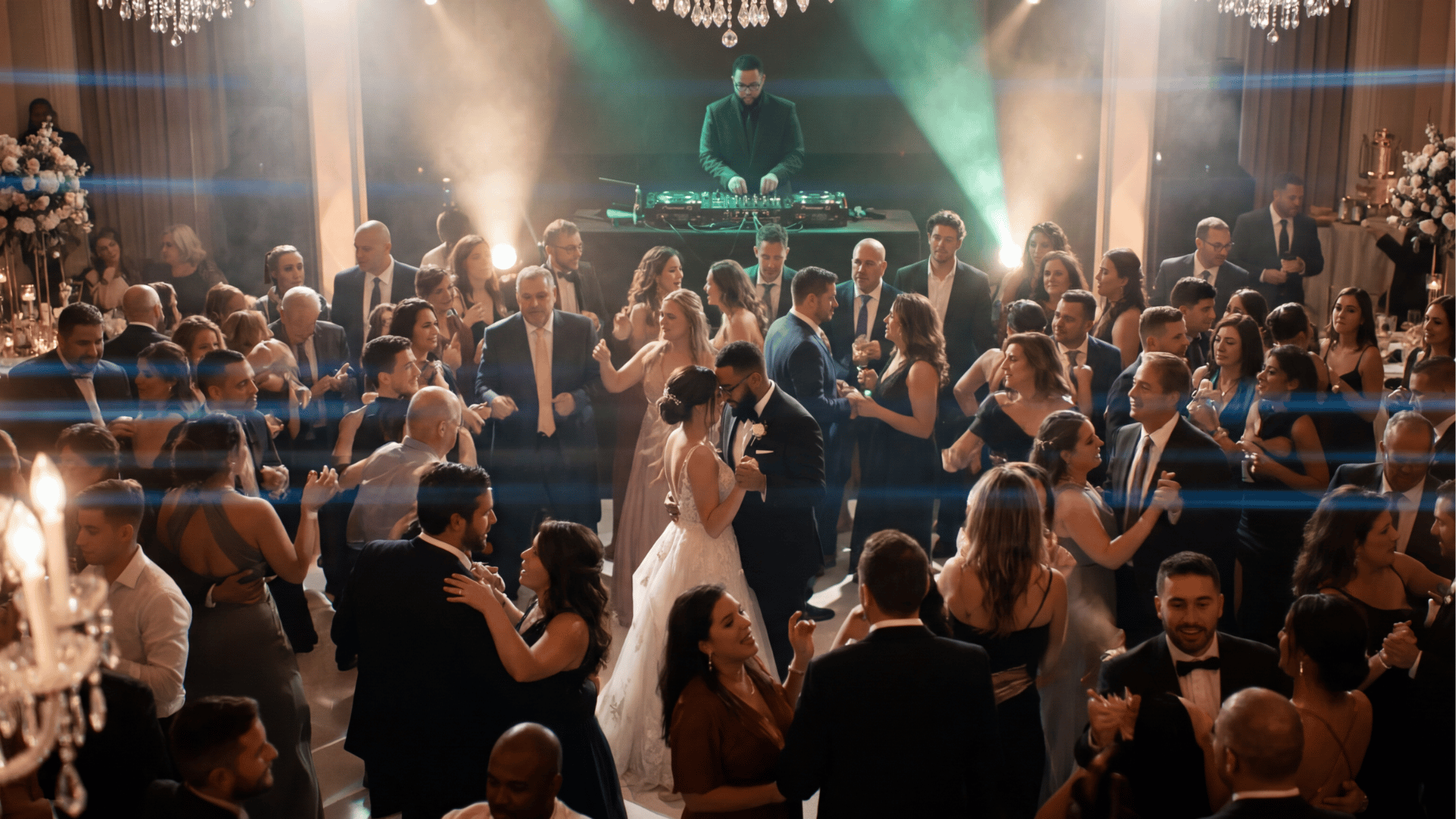 Bride and groom dancing surrounded by guests in formal attire at a wedding reception with DJ in background.