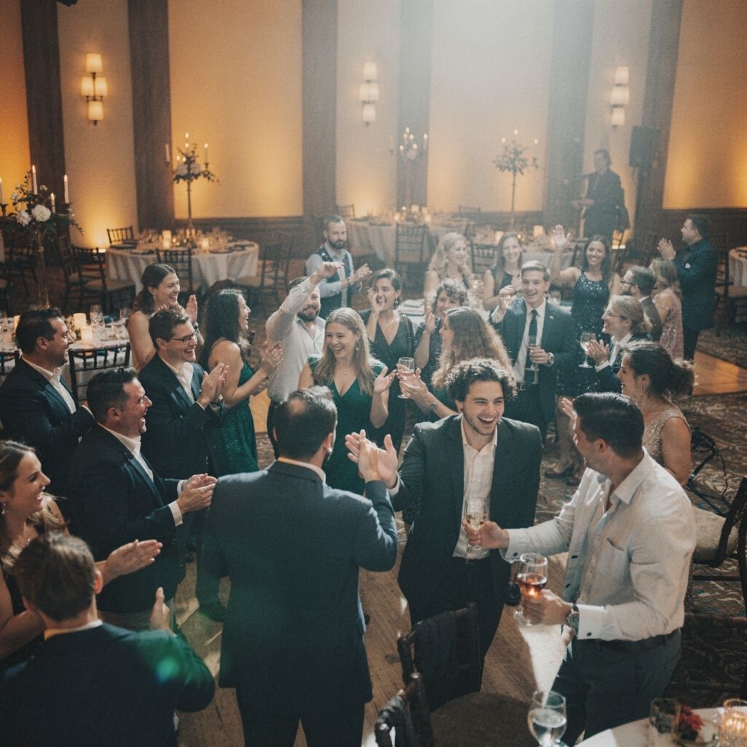 Group of people in formal attire clapping and celebrating at an indoor event with round tables.