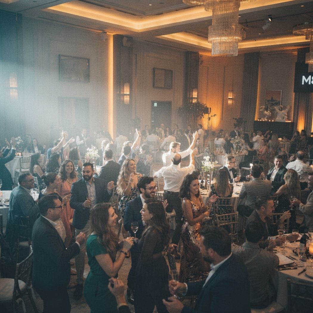 People dressed formally dancing and socializing at a warmly lit indoor event with tables and chandeliers.