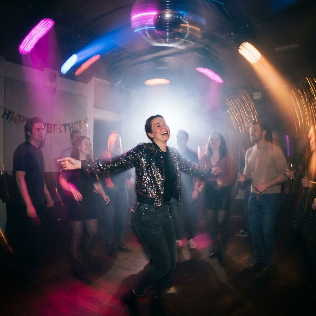 Person joyfully dancing with arms spread in a party room with colorful lights and guests watching.