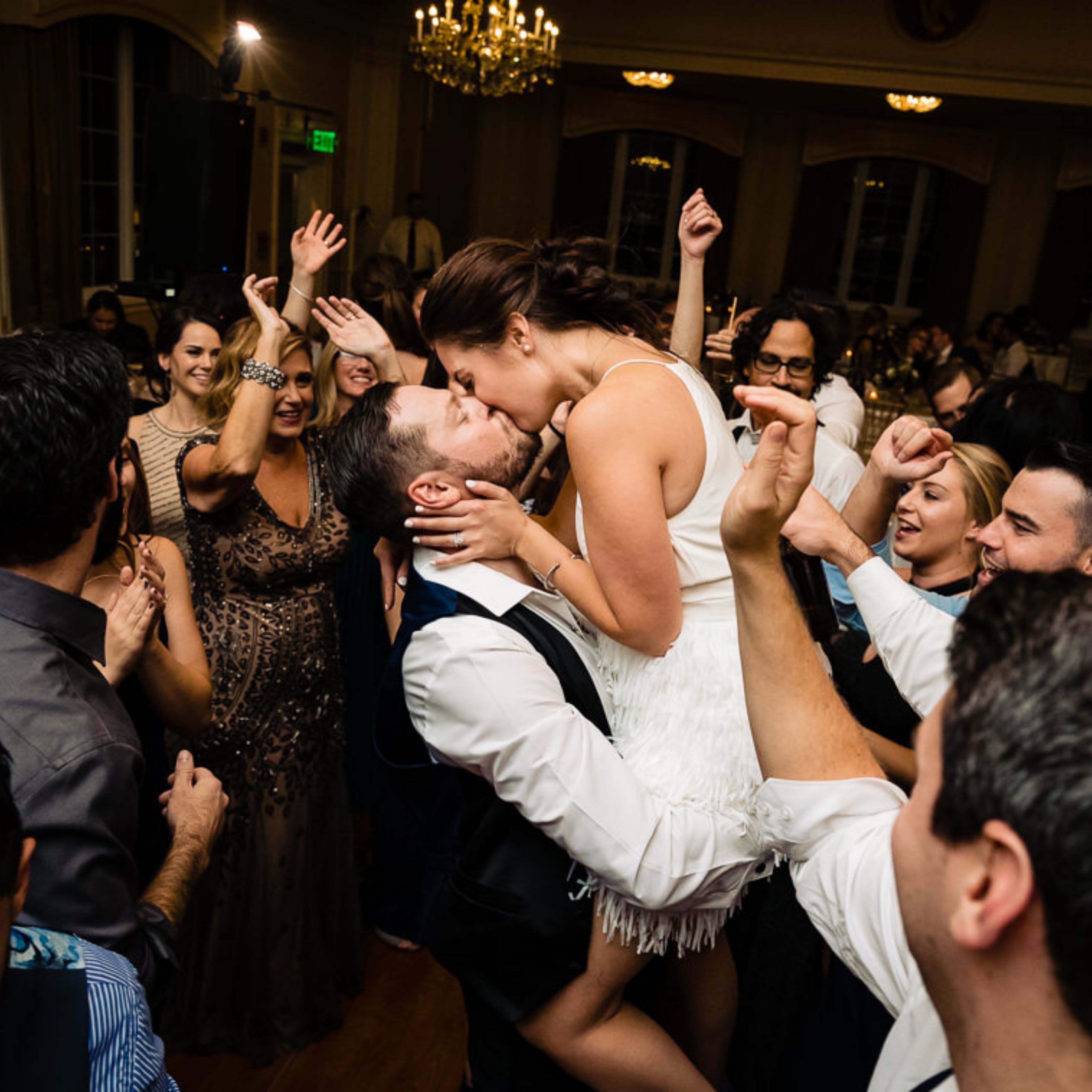 A couple kissing passionately while surrounded by dancing and cheering guests at a lively party.