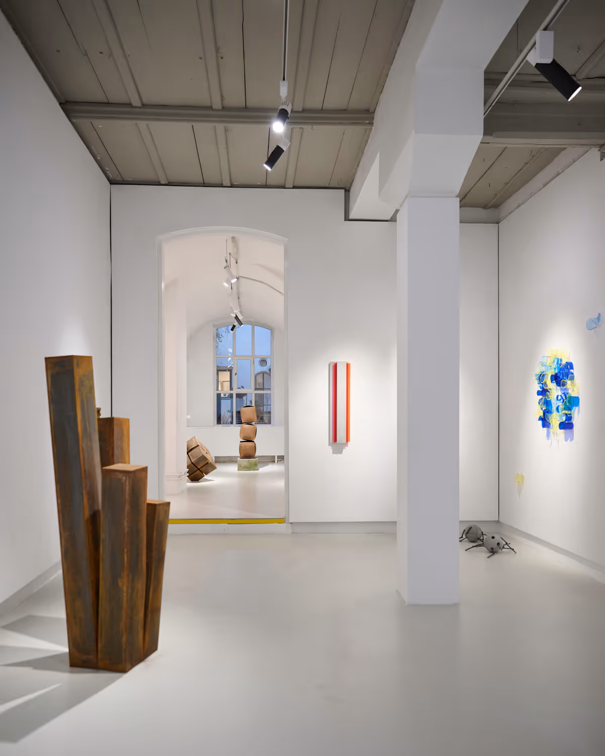 KunstKontor Nürnberg - Eine helle Kunstgalerie mit weißen Wänden, in der abstrakte Skulpturen und farbenfrohe Wandkunst ausgestellt sind, darunter hohe, rostfarbene Metallformen und übereinander gestapelte geometrische Figuren.