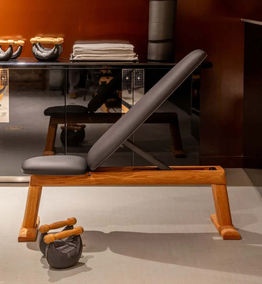 Banc de musculation en bois avec dossier rembourré incliné, accompagné de poids avec poignées en bois sur un sol clair.