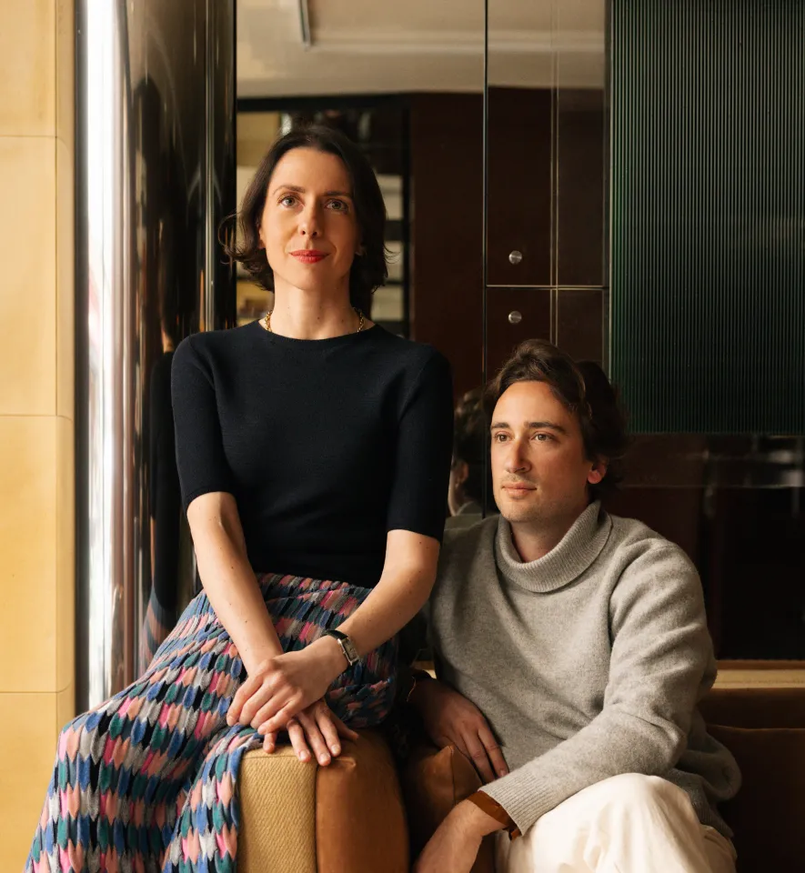A man sitting in a gray sweater and light pants alongside a woman standing in a black top and colored skirt.