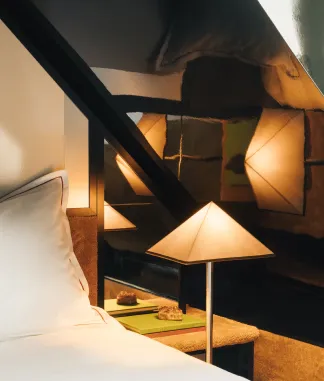 Triangular bedside lamp next to a white bed with a dark wall in the background.