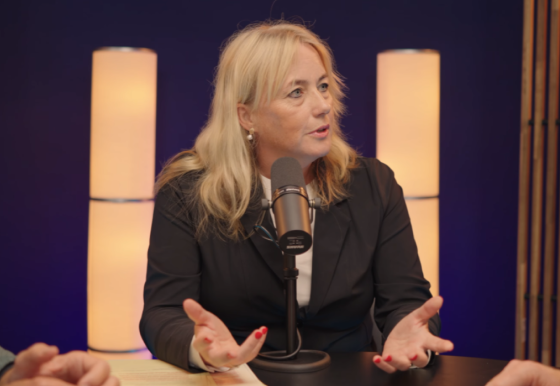 Blonde vrouw in zwarte jas spreekt in een microfoon tijdens een podcast of interview met zachte verlichting op de achtergrond.