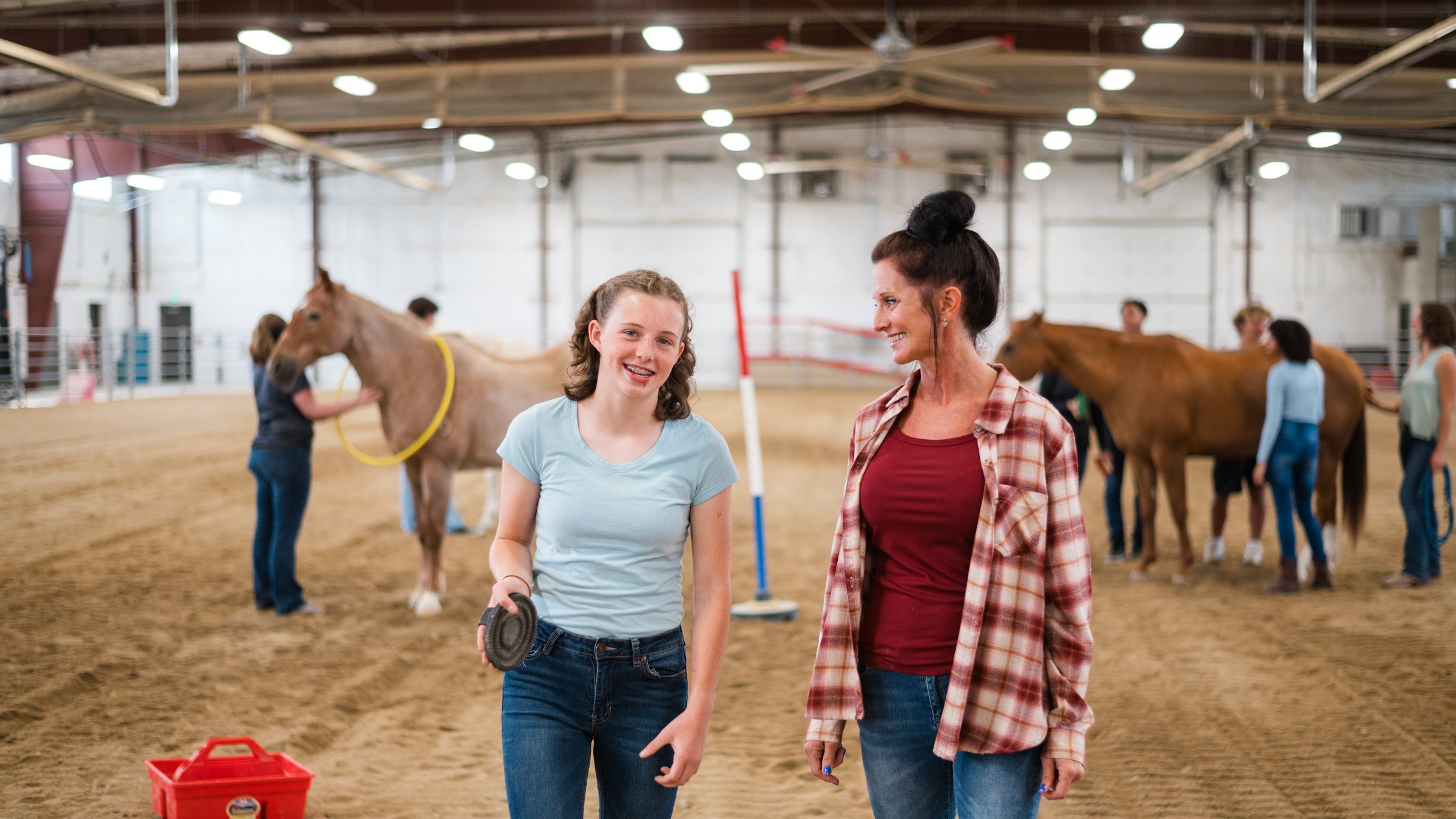 A youth and a caring adult walk together through an indoor arena during an equine-assisted therapy session at Idaho Youth Ranch, with horses and other participants in the background.