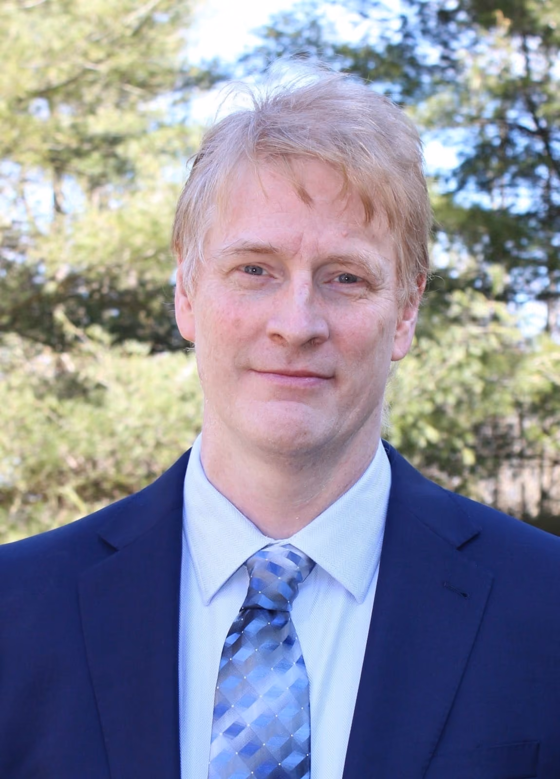 Portrait of a middle-aged man with light blond hair wearing a blue suit jacket, light blue dress shirt, and patterned blue tie, standing outdoors with green trees in the background.