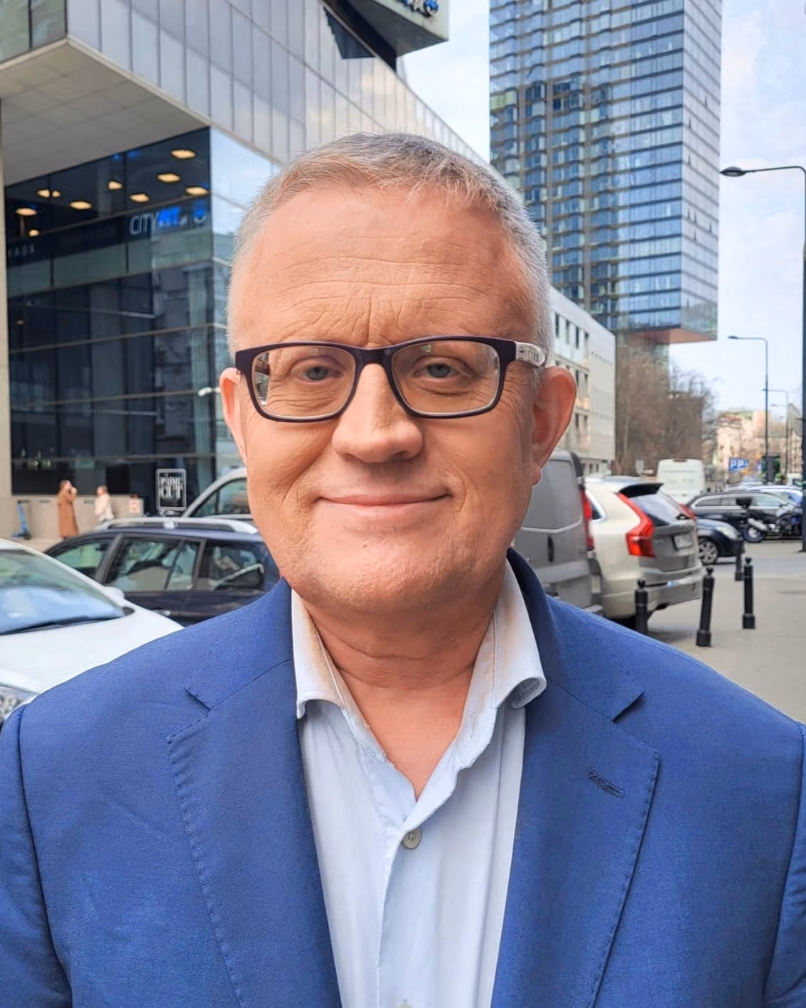 Middle-aged man wearing glasses, a light blue shirt, and a blue blazer, standing outdoors on a city street with modern buildings and parked cars in the background.