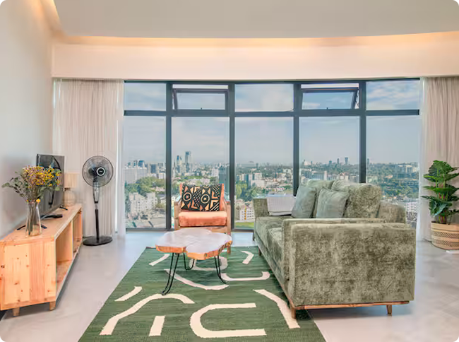 Modern living room with large windows overlooking a cityscape, featuring a green sofa, orange armchair, wooden coffee table, patterned rug, TV on wooden console, and standing fan.