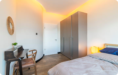 Modern bedroom with a bed, gray wardrobe, wooden desk with chair, and warm ambient lighting.