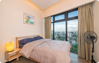 Bedroom with double bed, bedside table with lamp, standing fan, and large window overlooking a cityscape.