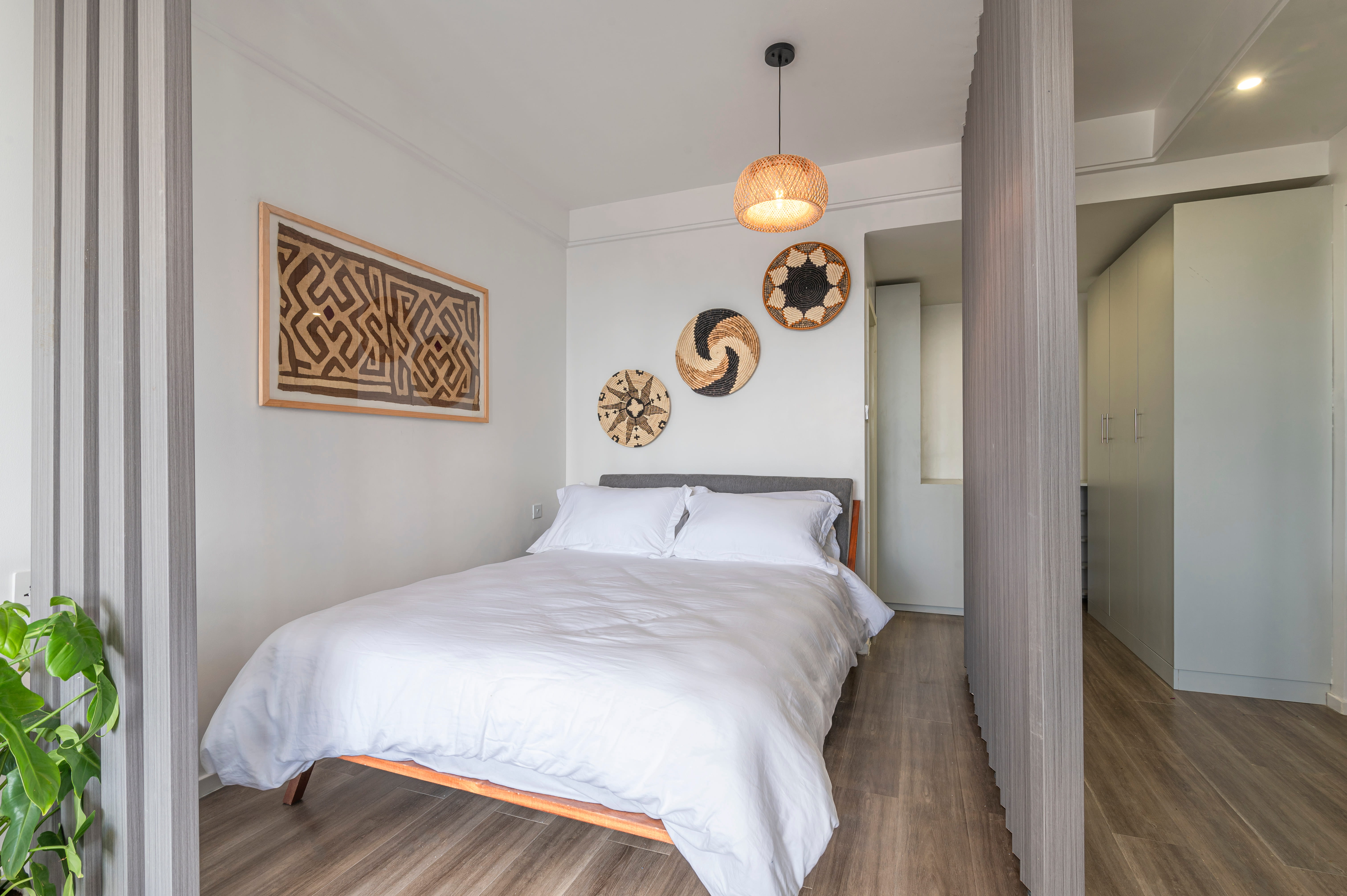 Minimalist bedroom with white bedding, wooden floor, wall decor, and a woven pendant light.