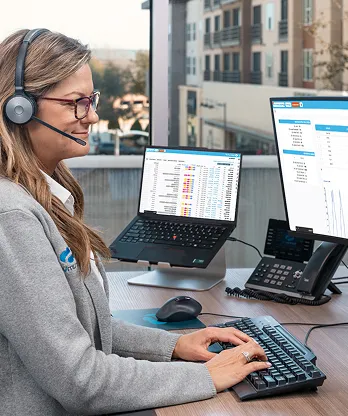 A woman wearing a headset works at a desk with two computer screens displaying spreadsheets.