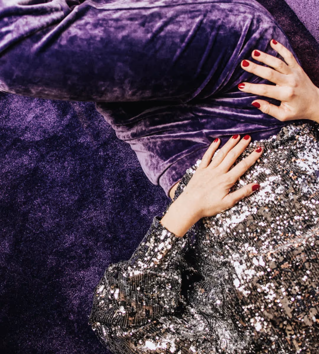 Gros plan de mains avec manucure rouge sur une jupe en velours violet et un haut à paillettes, tapis de l'Hôtel Château d’Eau 4 étoiles.