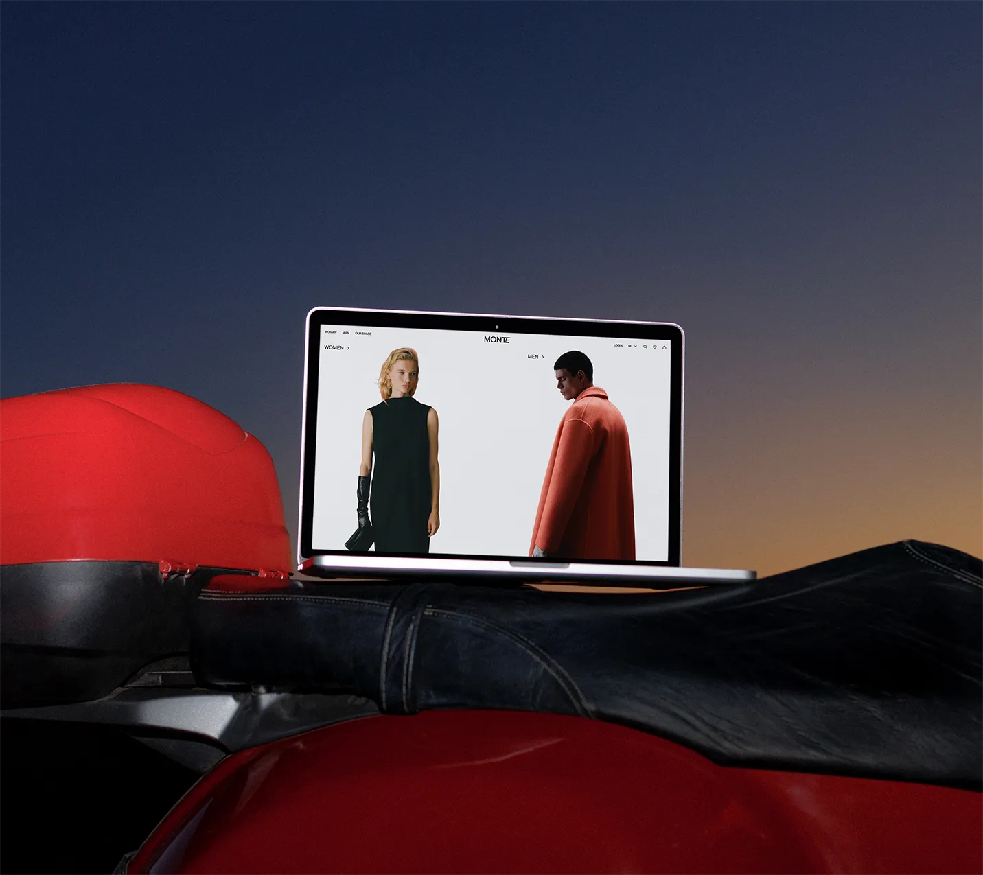 Open laptop displaying a fashion website with a woman in a black dress and a man in an orange coat on the screen, sitting on a red motorcycle seat against a twilight sky.