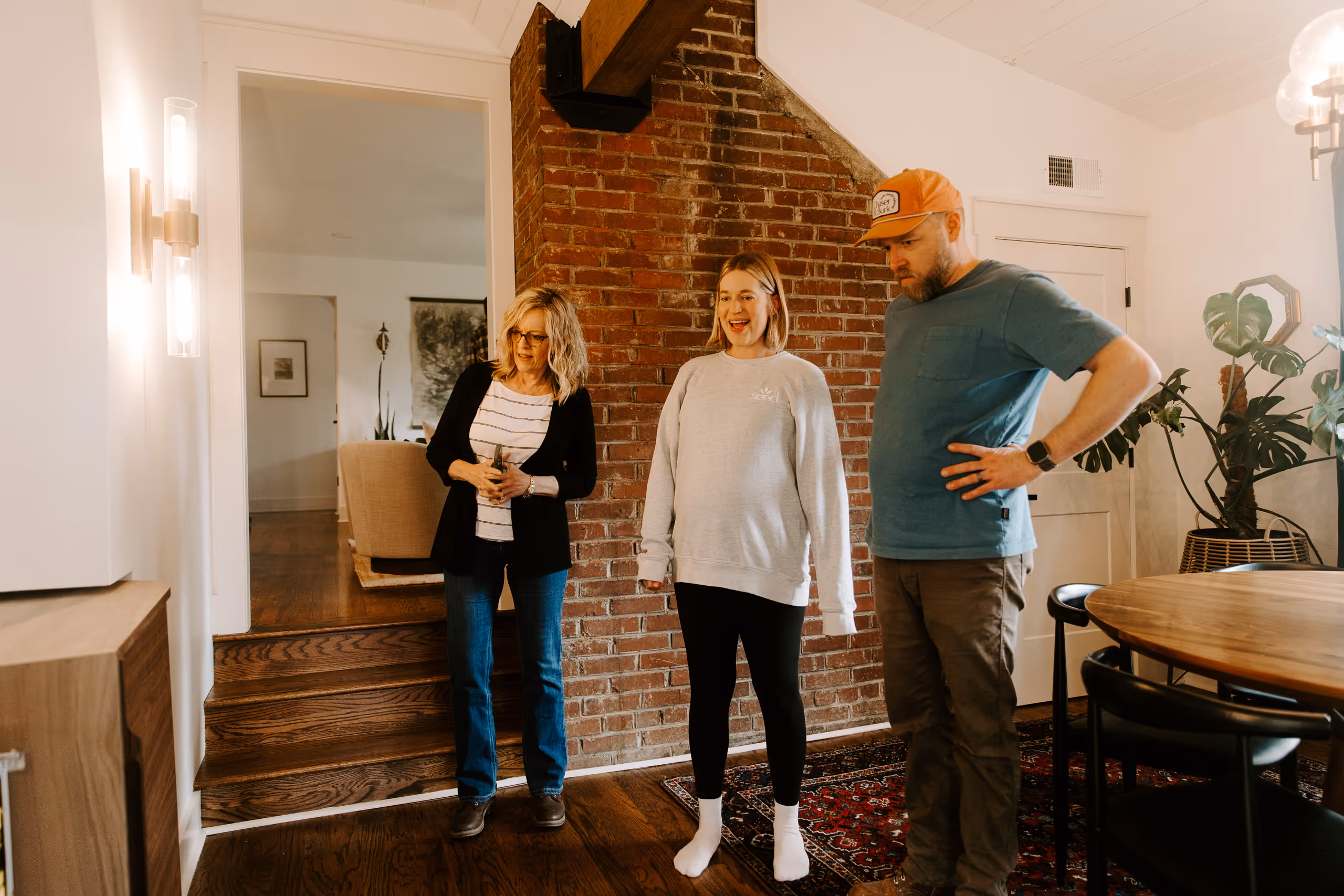 Three people standing in a room with a brick wall, wooden floor, and a round dining table, looking at something off-frame.