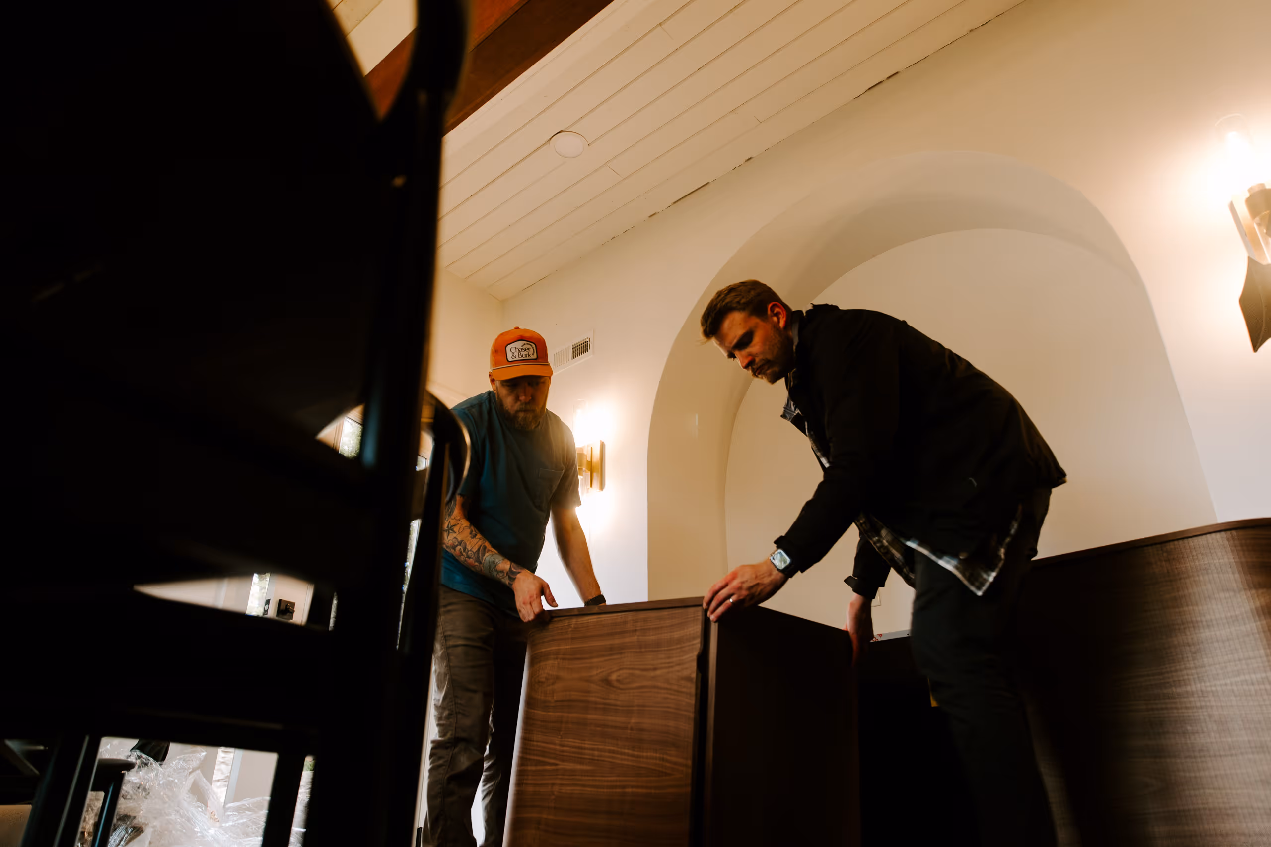 Two men assembling or moving a large wooden cabinet inside a room with arched wall and ceiling lights.