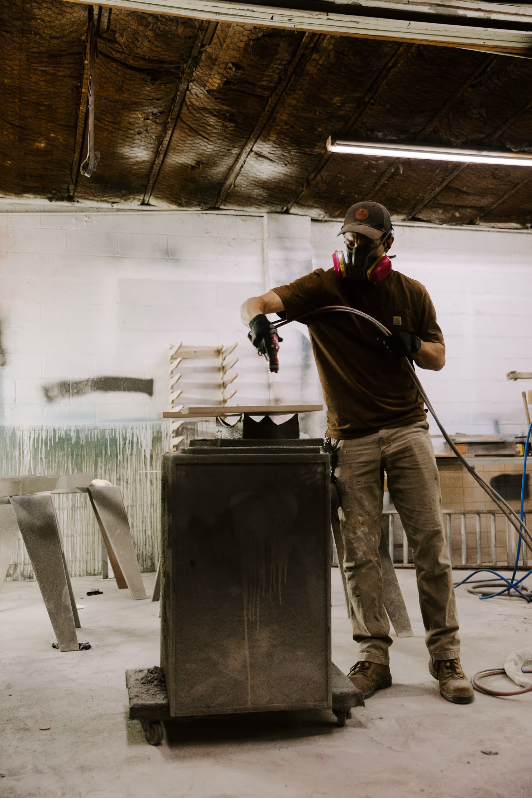Worker wearing a respirator mask and gloves spray painting a wooden object in a dusty workshop.