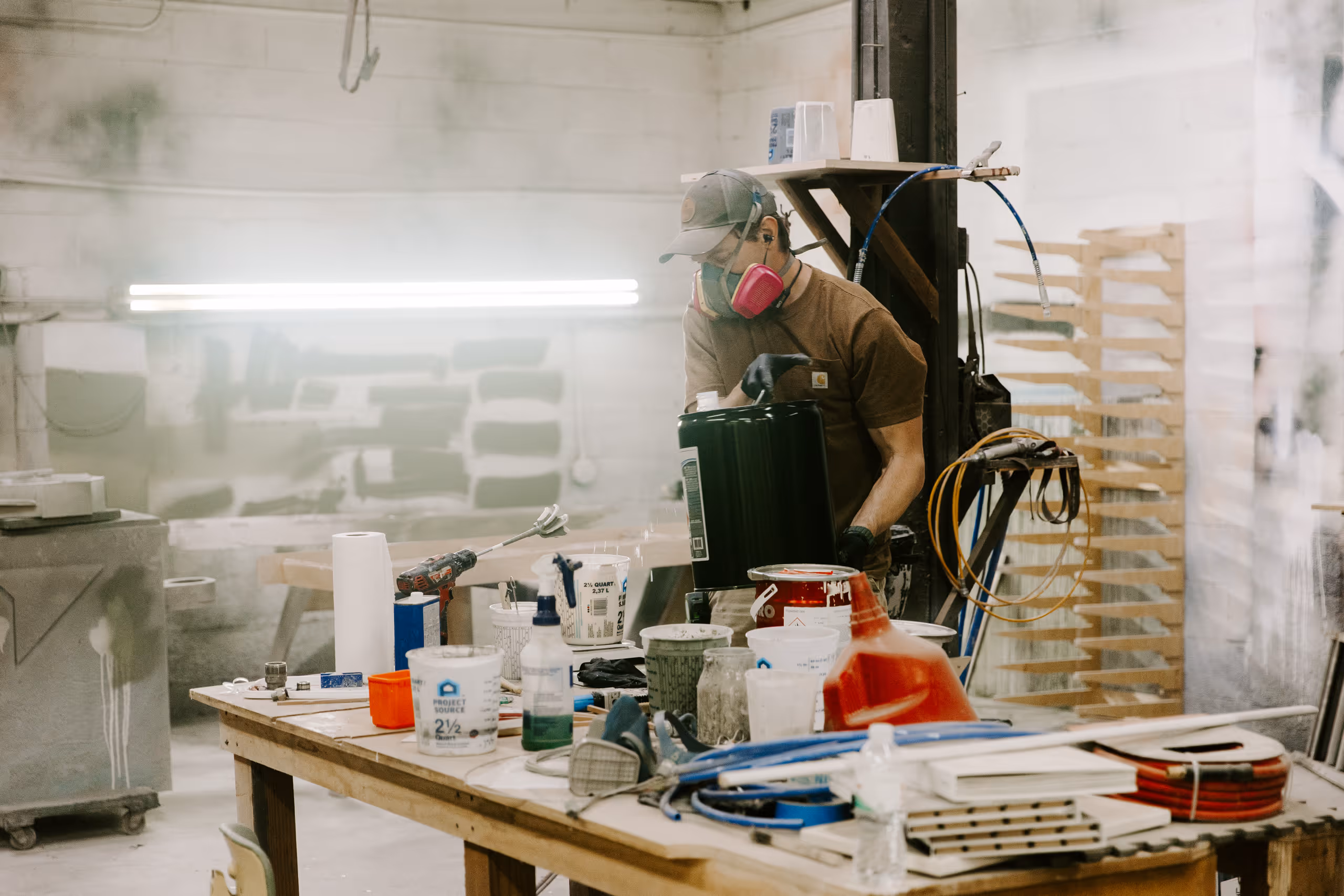 Man wearing a mask and gloves working with paint cans and equipment on a cluttered workshop table.
