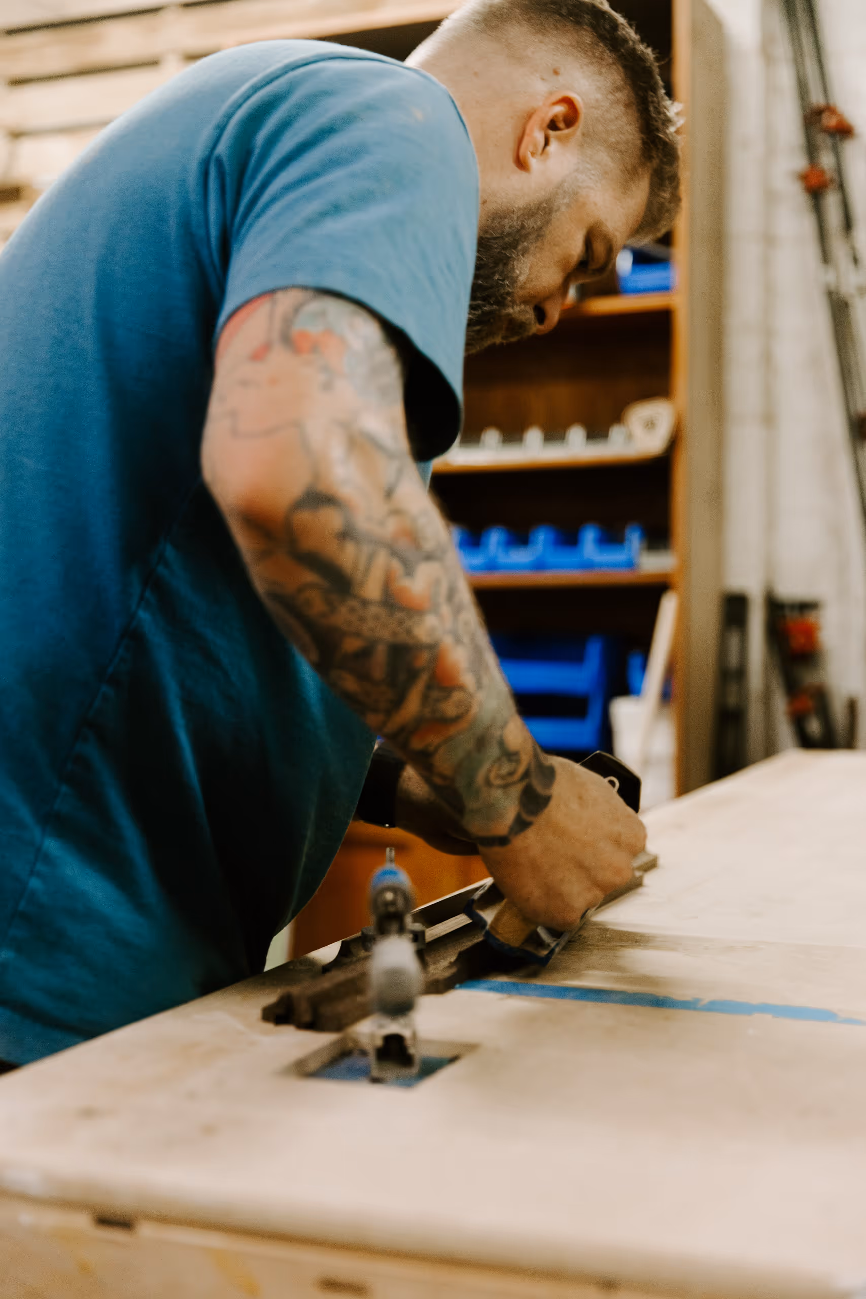 Tattooed man in a blue shirt working with a hand tool on a wooden surface in a workshop.