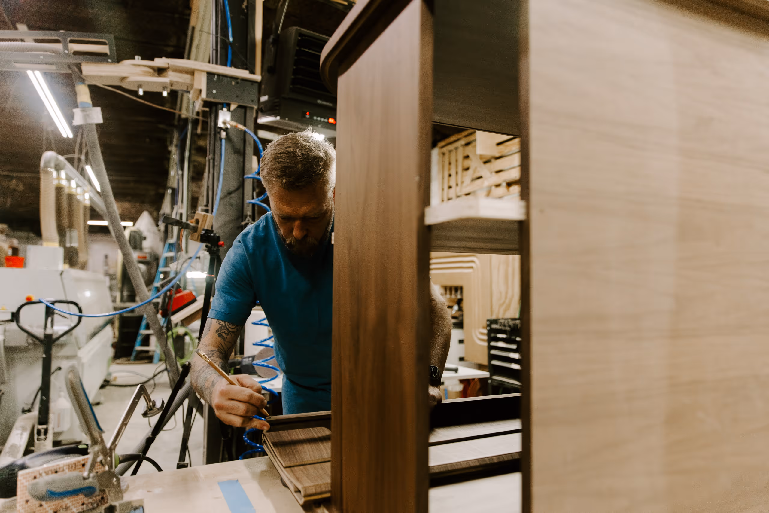 Man with tattoos working on wooden cabinet in a woodworking shop.