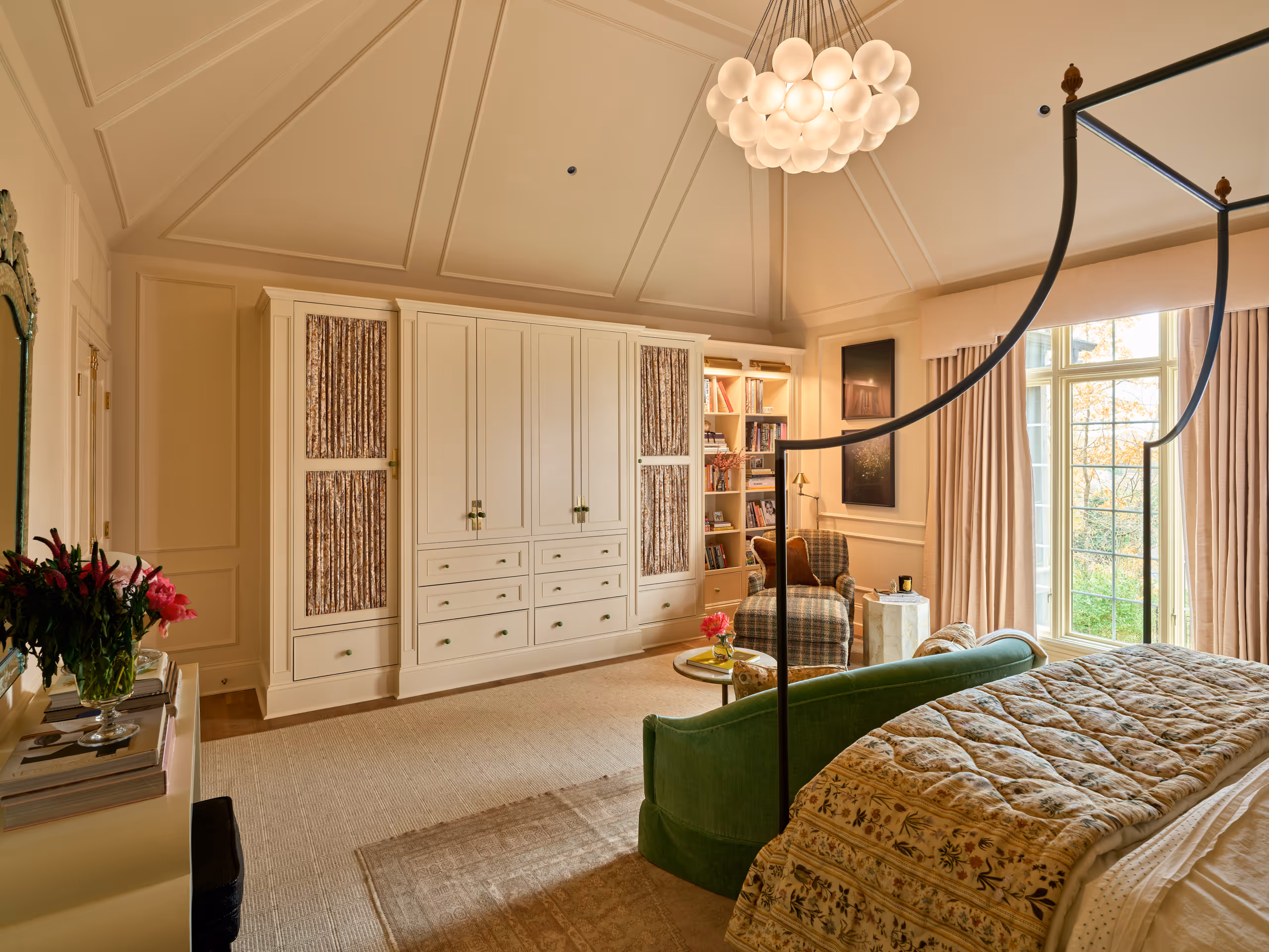 Elegant bedroom with a four-poster bed, green velvet bench, built-in cabinets with patterned curtains, armchair, and large window