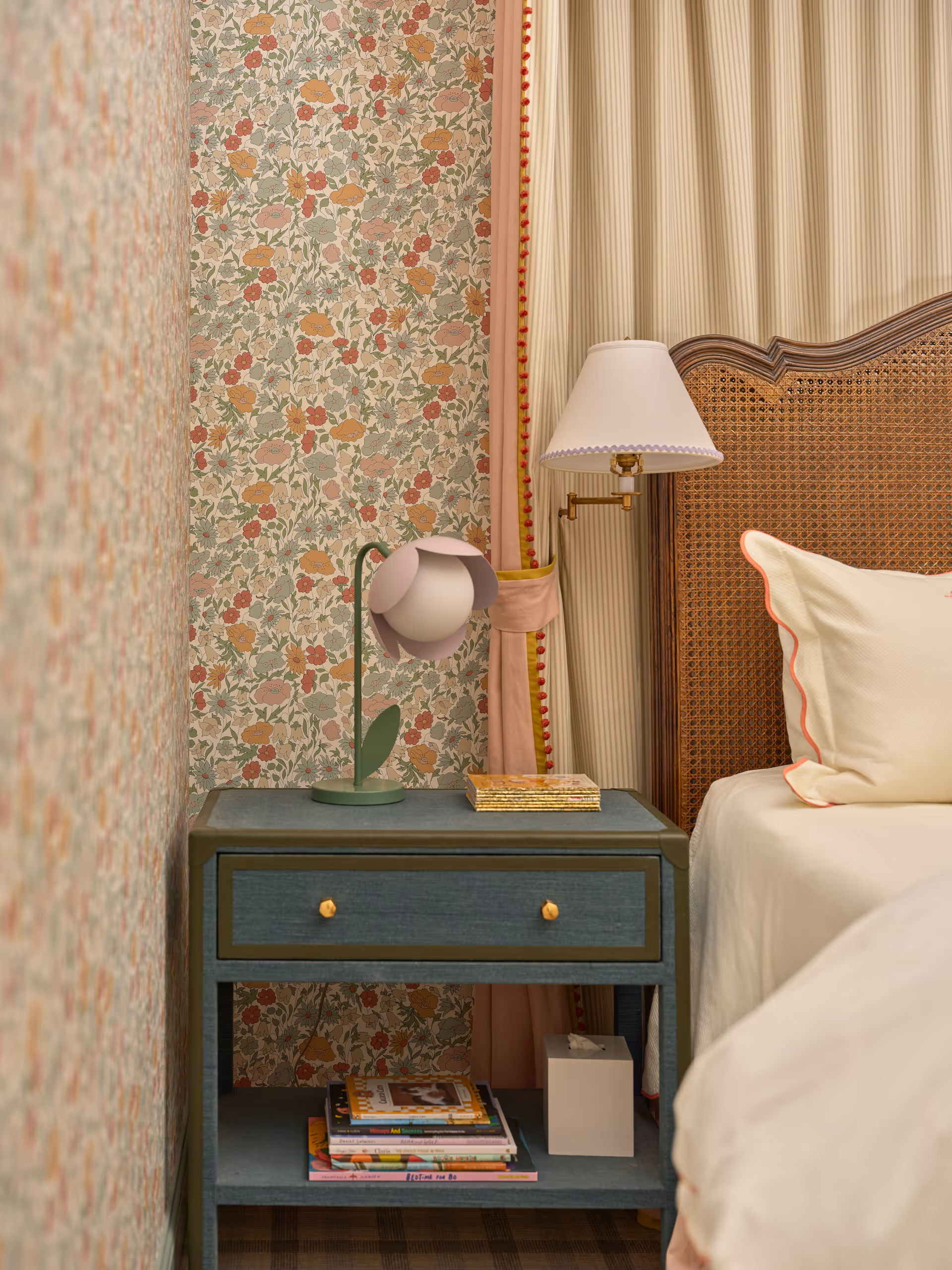 Blue bedside table with gold knobs holding a flower-shaped lamp, books, and a tissue box next to a wicker headboard bed.