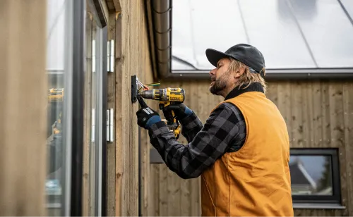 Man using a cordless drill to fix an electrical box on the outside wall of a wooden house.