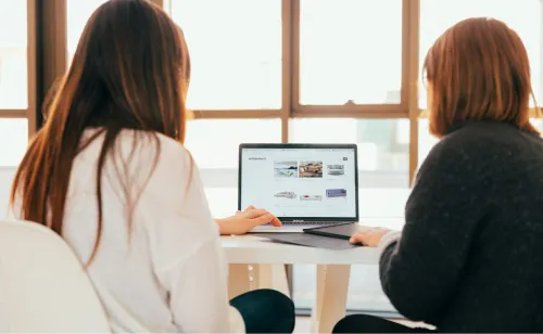Two people sitting at a table, looking at a laptop screen showing website images.