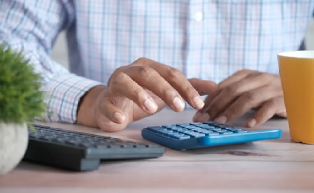 Person using a blue calculator at a desk with a keyboard, plant, and yellow mug nearby.