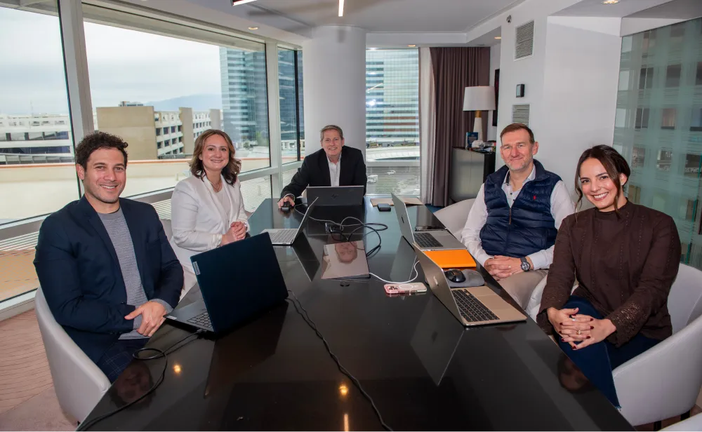 Five businesspeople seated around a conference table with laptops in a modern office with large windows.