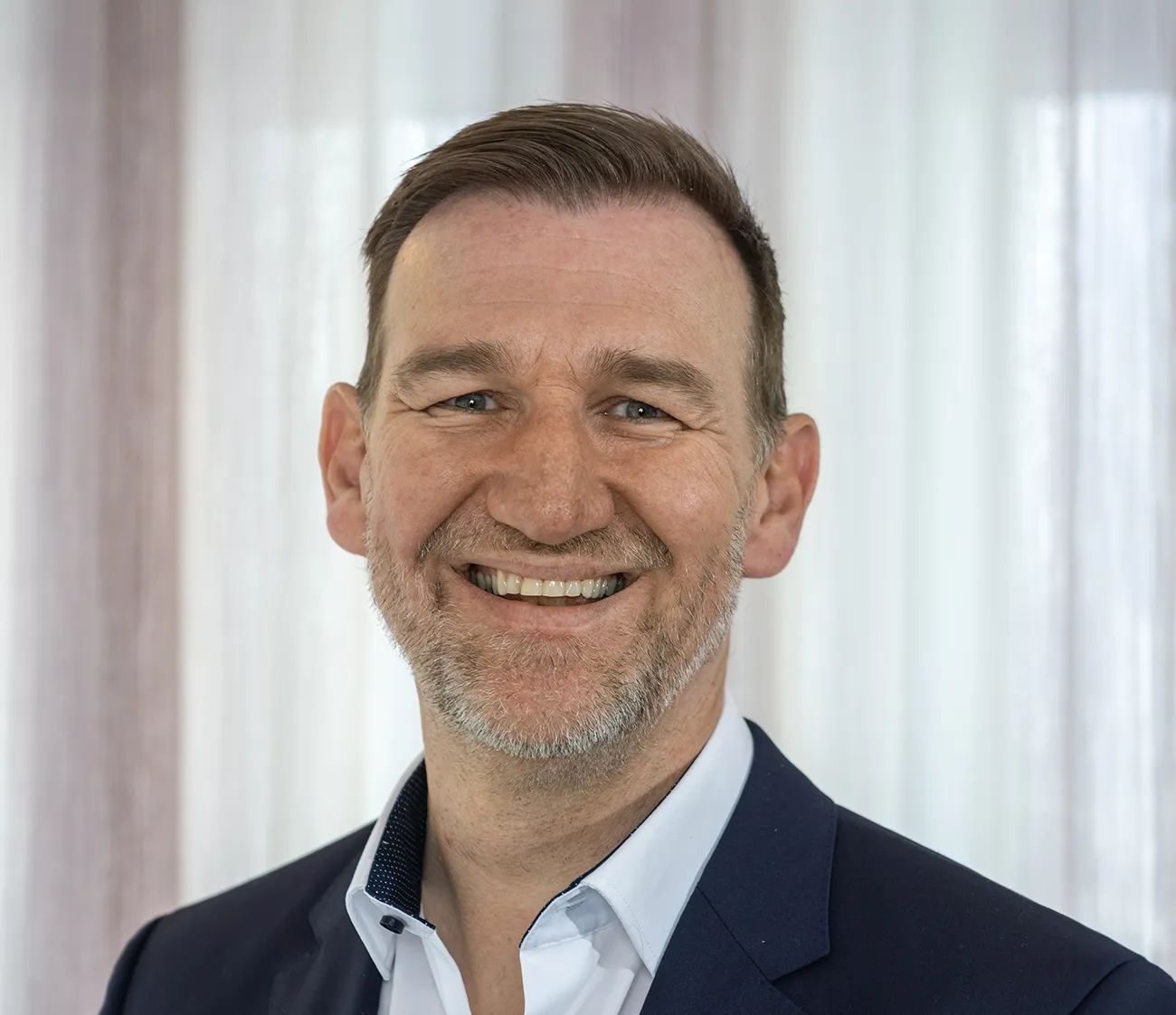 Smiling middle-aged man with beard wearing a blue suit jacket and light blue shirt in front of a blurred curtain.