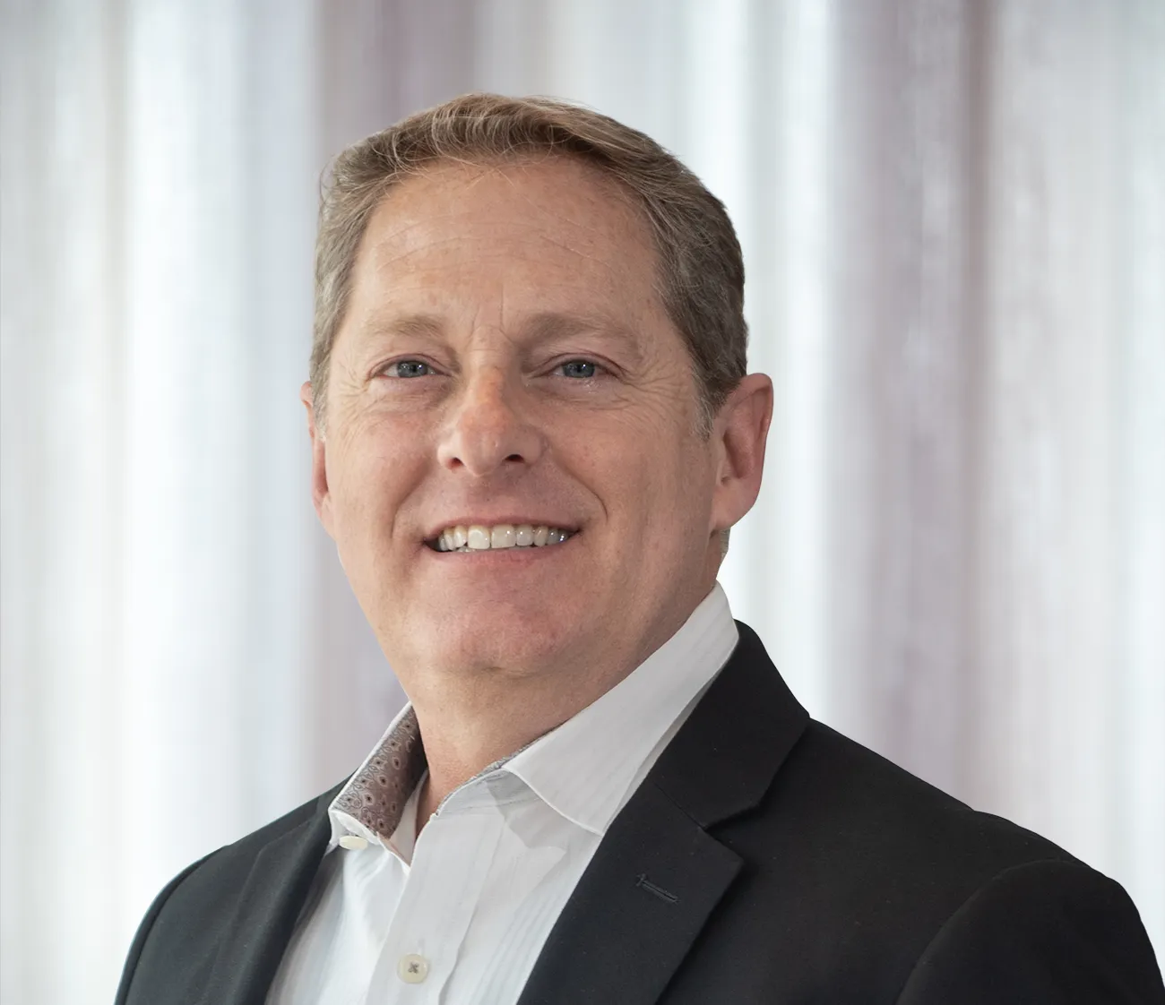 Smiling middle-aged man with light brown hair wearing a white shirt and black blazer.