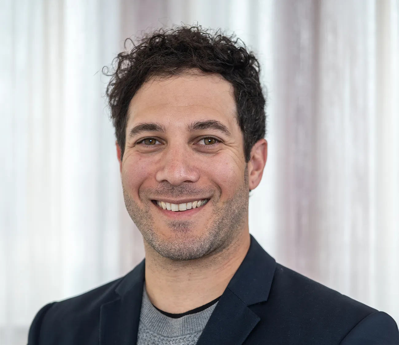 Smiling man with short curly hair, light beard, wearing a dark blazer and gray sweater.