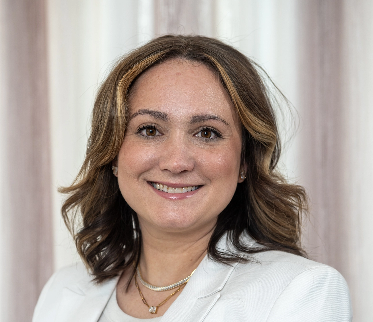 Smiling woman with brown eyes and wavy hair wearing a white blazer and gold necklaces.