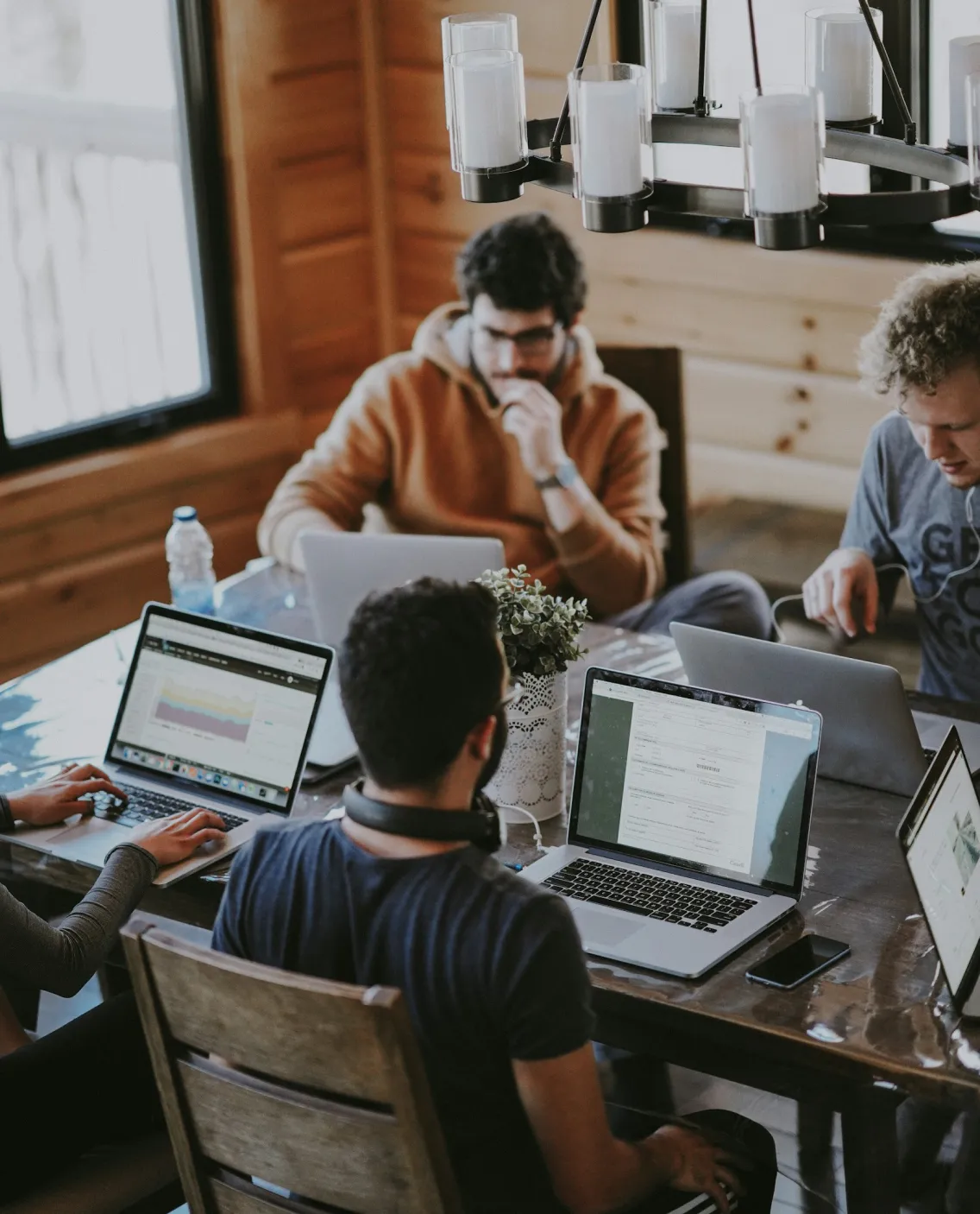 Four people working on laptops around a wooden table in a cozy, wood-paneled room with a chandelier.